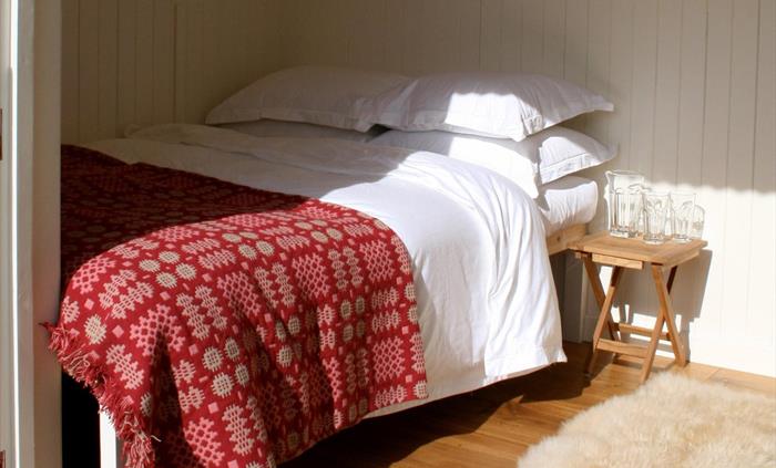 Double bed with the sun shining in with luxurious white cotton bed linen, a red traditional Welsh blanket and a sheepskin rug on the oak floor.